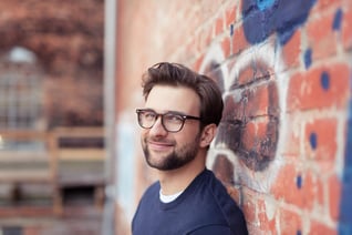 Portrait of Smiling Young Man with Facial Hair Wearing Eyeglasses and Leaning Against Brick Wall Painted with Graffiti-1_11zon